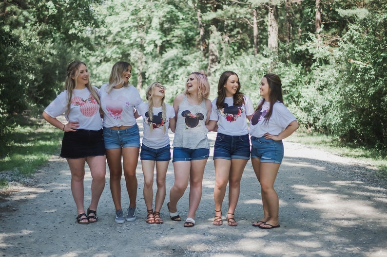 May include: Six friends wearing white t-shirts with a black and white Mickey Mouse head design and floral accents. They are standing on a dirt road with trees in the background.