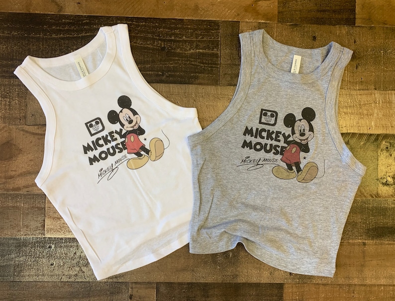 May include: Two cropped tank tops featuring a vintage-style Mickey Mouse graphic. One tank top is white with black text and the other is gray with black text. Both tank tops feature the text "Mickey Mouse" and a signature.