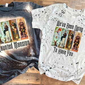May include: Two bleached t-shirts with a Haunted Mansion theme. The dark grey shirt has a graphic of the Haunted Mansion characters and the text "Haunted Mansion". The white shirt has a graphic of the Haunted Mansion characters and the text "We've Been Dying To Have You".