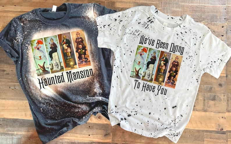 May include: Two bleached t-shirts with a Haunted Mansion theme. The dark grey shirt has a graphic of the Haunted Mansion characters and the text "Haunted Mansion". The white shirt has a graphic of the Haunted Mansion characters and the text "We've Been Dying To Have You".