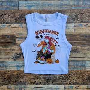 May include: White sleeveless crop top with a graphic print of a character from the movie "Nightmare Before Christmas". The graphic features the character Sally with Jack Skellington and Zero. The text "Nightmare on Main Street" is printed on the shirt.
