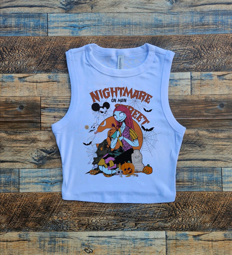 May include: White sleeveless crop top with a graphic print of a character from the movie "Nightmare Before Christmas". The graphic features the character Sally with Jack Skellington and Zero. The text "Nightmare on Main Street" is printed on the shirt.