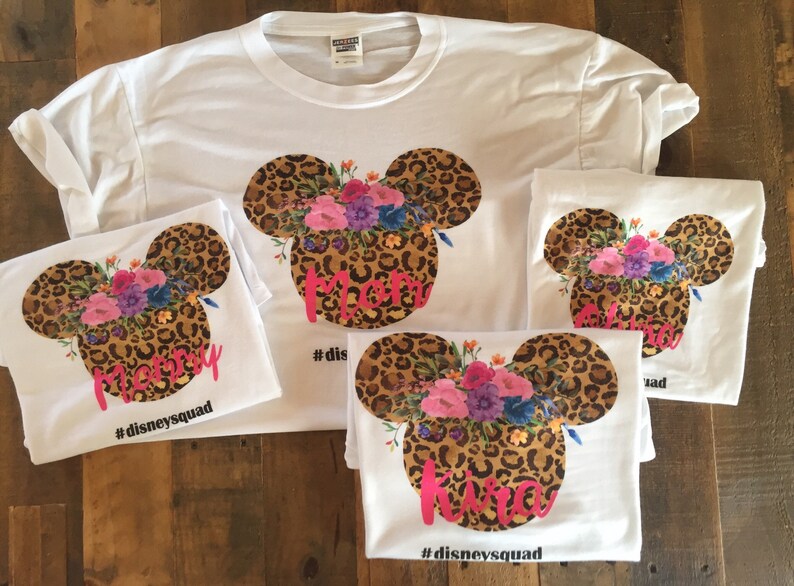 May include: Four white t-shirts with a leopard print Mickey Mouse head design with floral accents. The shirts have the words "Mommy", "Kira", and "#disneysquad" printed on them.