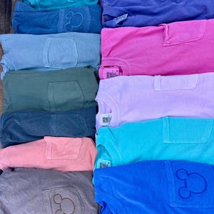 May include: A selection of colourful t-shirts in shades like teal, blue, pink, and purple. Each shirt has a pocket and a Mickey Mouse outline on the pocket. The shirts are neatly stacked, displaying a range of colours.
