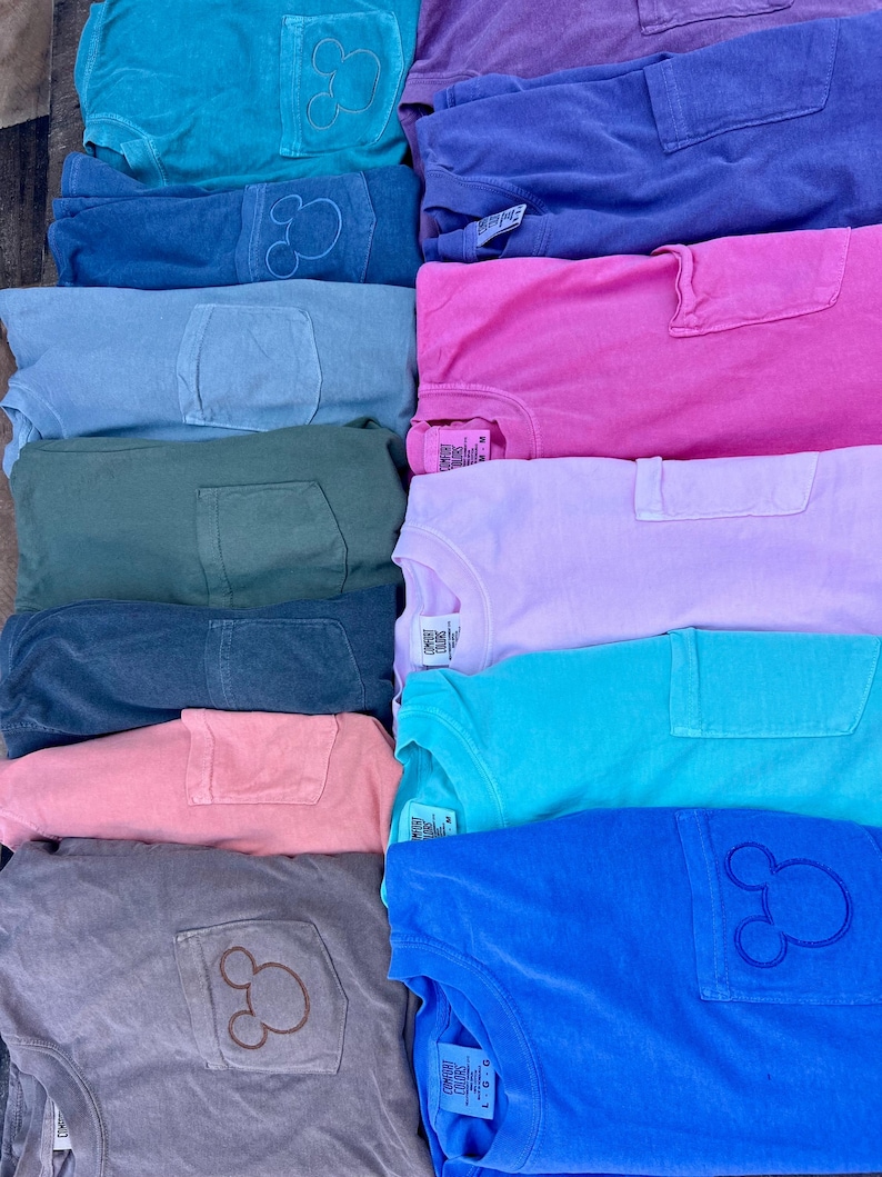 May include: A selection of colourful t-shirts in shades like teal, blue, pink, and purple. Each shirt has a pocket and a Mickey Mouse outline on the pocket. The shirts are neatly stacked, displaying a range of colours.