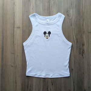 May include: A light blue tank top with a Mickey Mouse graphic. The sleeveless top features a round neckline and a small embroidered Mickey Mouse face on the chest. The tank top is made of a soft, lightweight material.