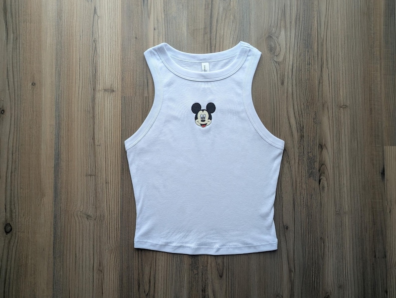 May include: A light blue tank top with a Mickey Mouse graphic. The sleeveless top features a round neckline and a small embroidered Mickey Mouse face on the chest. The tank top is made of a soft, lightweight material.