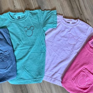 May include: Four short-sleeved t-shirts in shades of blue, teal, lavender, and pink. Each shirt features a Mickey Mouse outline on the chest and a pocket with the same design. The shirts are laid out on a wooden surface.