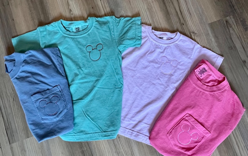 May include: Four short-sleeved t-shirts in shades of blue, teal, lavender, and pink. Each shirt features a Mickey Mouse outline on the chest and a pocket with the same design. The shirts are laid out on a wooden surface.