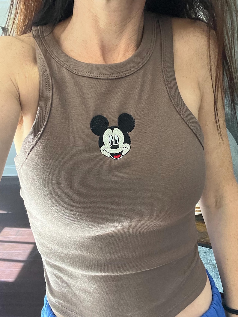 May include: A brown tank top with a round neckline and a small embroidered image of Mickey Mouse on the front. The sleeveless top is made of a soft, ribbed fabric.