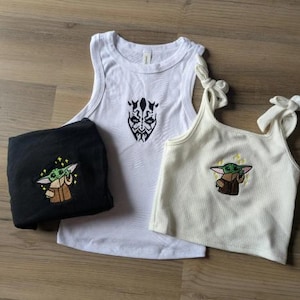 May include: A white tank top with a black Darth Maul embroidery, a folded black shirt with a green Grogu embroidery, and a cream-colored ribbed tank top with tie-shoulder straps and a green Grogu embroidery featuring small yellow stars.