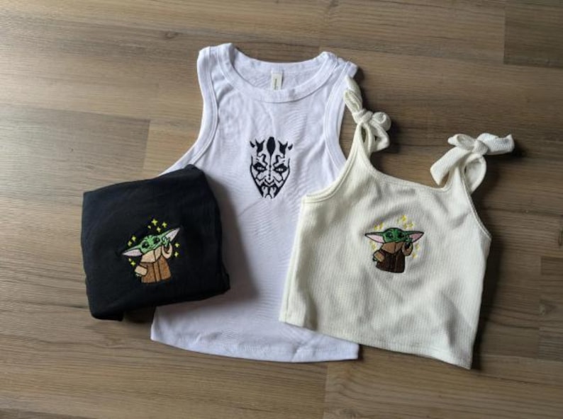 May include: A white tank top with a black Darth Maul embroidery, a folded black shirt with a green Grogu embroidery, and a cream-colored ribbed tank top with tie-shoulder straps and a green Grogu embroidery featuring small yellow stars.