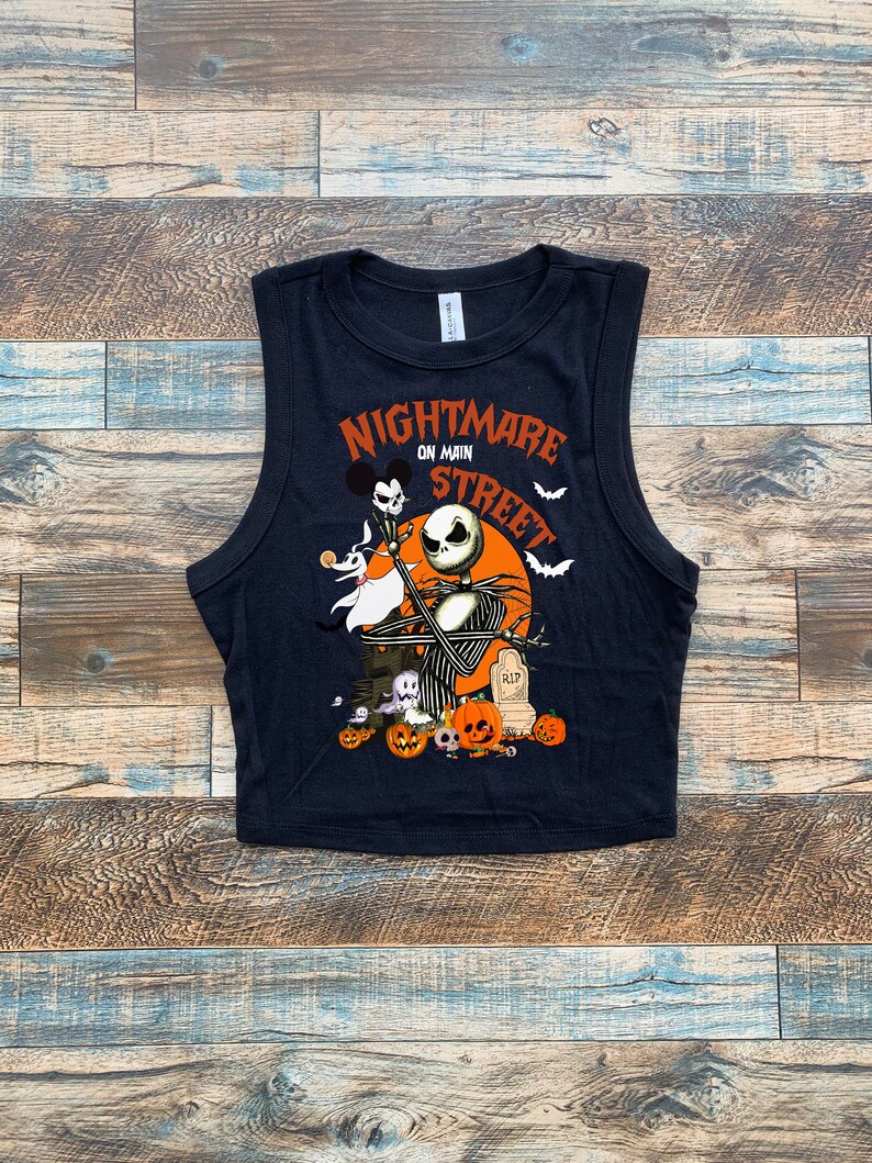 May include: Black sleeveless crop top with a graphic print of Jack Skellington from the Nightmare Before Christmas movie. The graphic includes the text "Nightmare on Main Street" and features Jack Skellington holding a microphone with Zero the ghost dog by his side. The graphic also includes pumpkins, a tombstone, and bats.