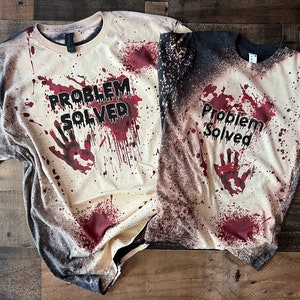 Pode incluir: Duas camisetas de manga curta com um efeito desbotado e desgastado em tons de cinza escuro e creme. Ambas as camisetas exibem um padrão de respingos vermelhos que lembra sangue, uma marca de mão vermelha e o texto preto "PROBLEM SOLVED".