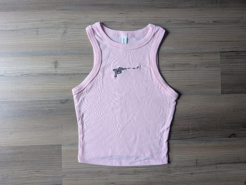 May include: A light pink sleeveless tank top with a crew neckline and ribbed trim. Centered on the chest is a small, black embroidered graphic of a ray gun firing a projectile with the text "pew pew" written in a simple, lowercase font.