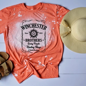 K&ouml;nnte beinhalten: Orangefarbenes T-Shirt mit einem Design aus Bleichmittelspritzern, das eine schwarz-wei&szlig;e Grafik eines Etiketts mit der Aufschrift "Winchester Brothers Since 1983 Saving People Hunting Things A Family Business" zeigt.