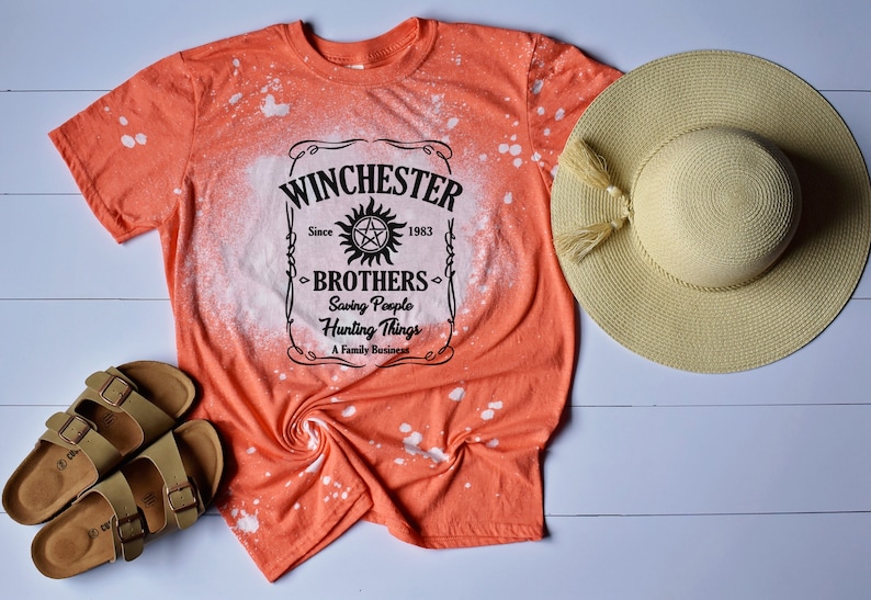 K&ouml;nnte beinhalten: Orangefarbenes T-Shirt mit einem Design aus Bleichmittelspritzern, das eine schwarz-wei&szlig;e Grafik eines Etiketts mit der Aufschrift "Winchester Brothers Since 1983 Saving People Hunting Things A Family Business" zeigt.