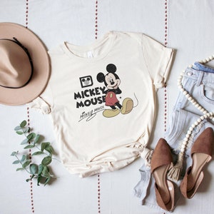 May include: A cream colored t-shirt with a vintage Mickey Mouse graphic. The graphic features Mickey Mouse in black and red with the text "Mickey Mouse" in black. The shirt is laid out on a white surface with a brown hat, blue jeans, and a wooden bead necklace.