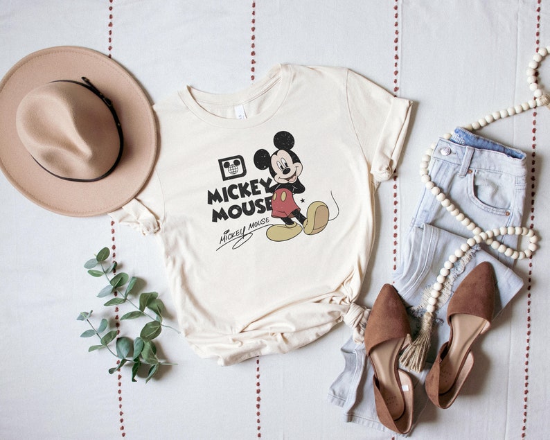 May include: A cream colored t-shirt with a vintage Mickey Mouse graphic. The graphic features Mickey Mouse in black and red with the text "Mickey Mouse" in black. The shirt is laid out on a white surface with a brown hat, blue jeans, and a wooden bead necklace.