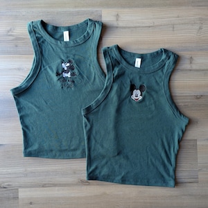 May include: Two forest green tank tops. One top features an embroidered Mickey Mouse design with the year "1928" below. The other top has an embroidered Mickey Mouse face. Both tops have a rounded neckline and sleeveless design.