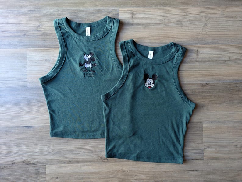 May include: Two forest green tank tops. One top features an embroidered Mickey Mouse design with the year "1928" below. The other top has an embroidered Mickey Mouse face. Both tops have a rounded neckline and sleeveless design.