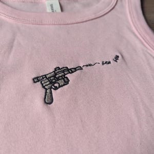 May include: A light pink tank top featuring a black embroidered graphic of a ray gun. The design includes a wavy line extending from the barrel of the gun, followed by the embroidered text "pew pew" in a small, simple, black cursive font.