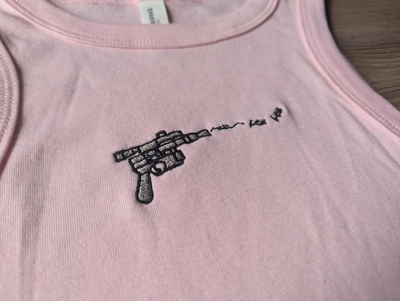 May include: A light pink tank top featuring a black embroidered graphic of a ray gun. The design includes a wavy line extending from the barrel of the gun, followed by the embroidered text "pew pew" in a small, simple, black cursive font.
