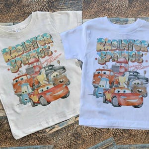 May include: Two white t-shirts with a graphic print of the Cars characters from the movie Cars. The graphic features the characters in front of a sign that says "Radiator Springs" with the text "Welcome to Radiator Springs" underneath.