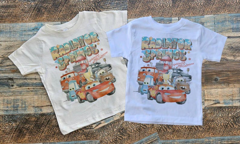 May include: Two white t-shirts with a graphic print of the Cars characters from the movie Cars. The graphic features the characters in front of a sign that says "Radiator Springs" with the text "Welcome to Radiator Springs" underneath.