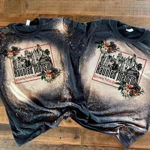 May include: Two black t-shirts with a bleach-splattered design featuring a black and white illustration of the Haunted Mansion with the text "Haunted Mansion Welcoming Foolish Mortals" and floral accents.