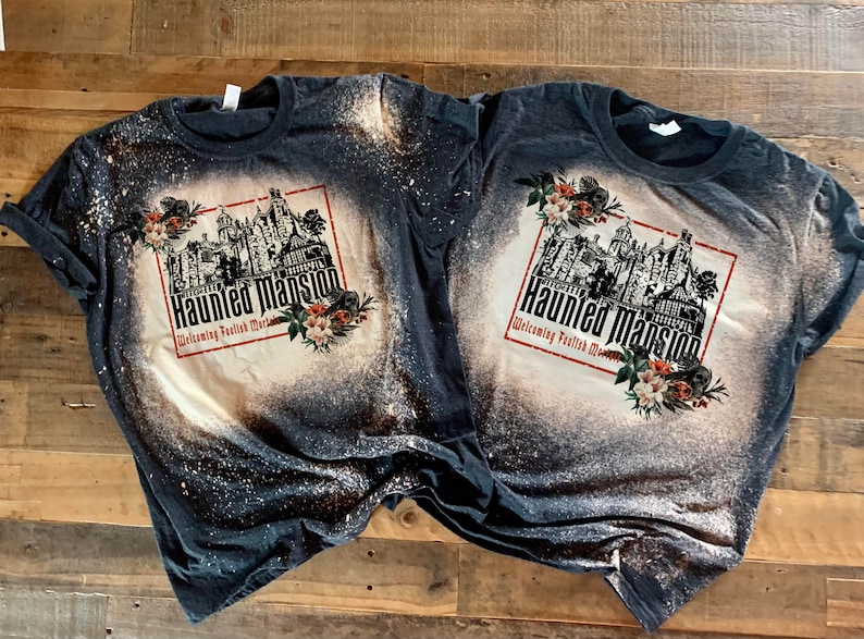 May include: Two black t-shirts with a bleach-splattered design featuring a black and white illustration of the Haunted Mansion with the text "Haunted Mansion Welcoming Foolish Mortals" and floral accents.