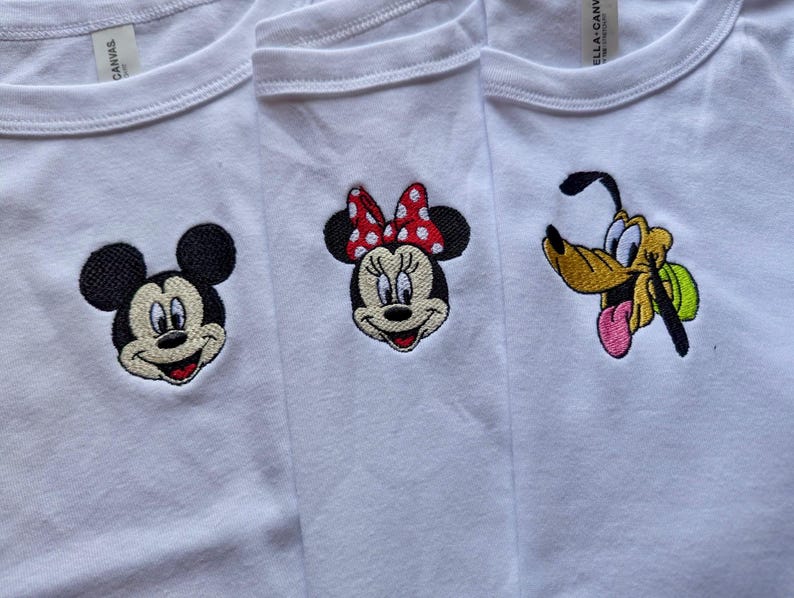 May include: Three white t-shirts with embroidered cartoon character faces. Mickey Mouse, Minnie Mouse with a red polka dot bow, and Pluto are featured. The shirts are made of a soft material.