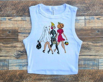 May include: White sleeveless crop top with a graphic print of three women dressed as a ghost, skeleton, and pumpkin for Halloween. The women are carrying trick-or-treat bags.