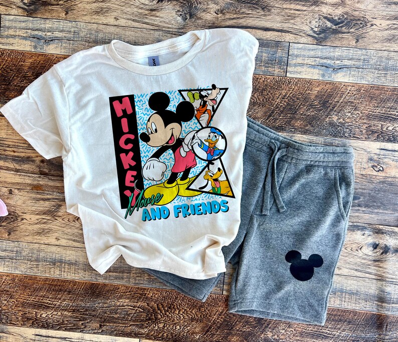 Puede incluir: Una camiseta blanca con un gr&aacute;fico vintage de Mickey Mouse que presenta a Mickey Mouse y sus amigos. El gr&aacute;fico est&aacute; en un estilo colorido de los 90 con el texto "Mickey Mouse and Friends". La camiseta se combina con pantalones de ch&aacute;ndal grises con un gr&aacute;fico de cabeza de Mickey Mouse negro.