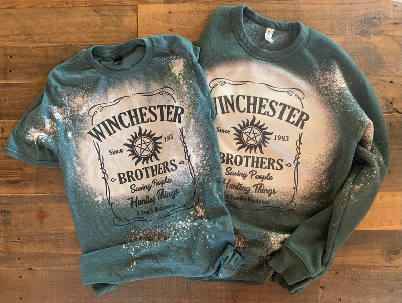 K&ouml;nnte beinhalten: Zwei gr&uuml;ne Shirts mit Bleiche-Spritzer-Designs. Die Shirts zeigen den Text "Winchester Brothers" mit einem Symbol und den Text "Saving People Hunting Things A Family Business".