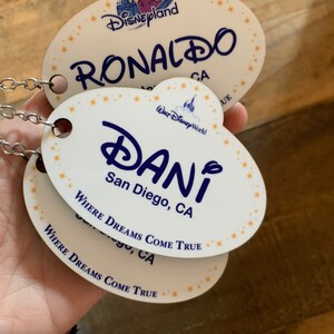 May include: Two white oval-shaped name tags with blue text and gold stars. The top tag reads "Disneyland RONALDO CA" and the bottom tag reads "WALT Disney World DANI San Diego, CA WHERE DREAMS COME TRUE".