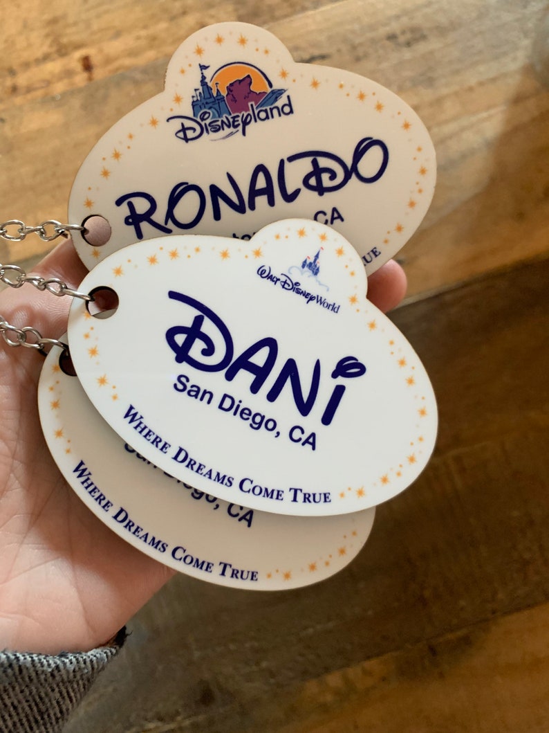 May include: Two white oval-shaped name tags with blue text and gold stars. The top tag reads "Disneyland RONALDO CA" and the bottom tag reads "WALT Disney World DANI San Diego, CA WHERE DREAMS COME TRUE".