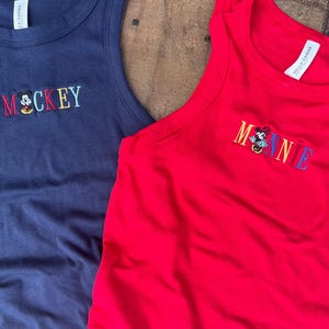 May include: Two sleeveless tank tops. One is navy blue with the word "MOCKEY" embroidered in colorful thread, with a cartoon character. The other is red with "MINNIE" embroidered in colorful thread, with a cartoon character.