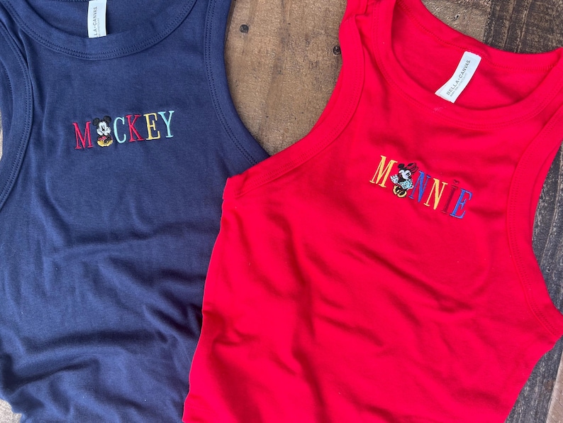 May include: Two sleeveless tank tops. One is navy blue with the word "MOCKEY" embroidered in colorful thread, with a cartoon character. The other is red with "MINNIE" embroidered in colorful thread, with a cartoon character.