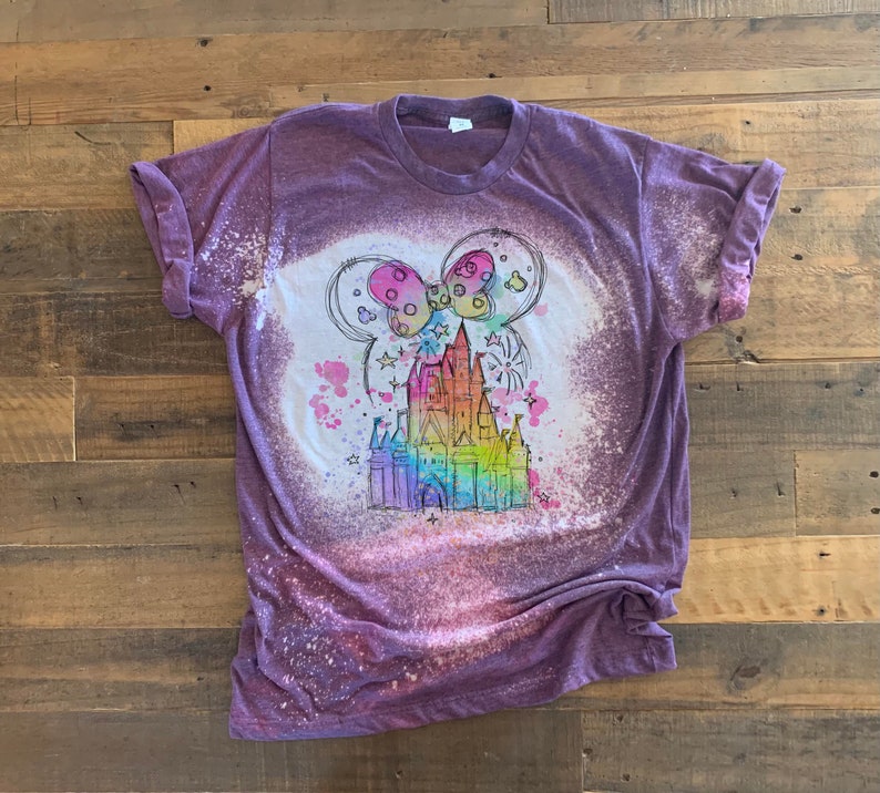 May include: Purple t-shirt with a bleach-splattered design featuring a rainbow-colored drawing of Cinderella's castle with a pink bow and Mickey Mouse ears.