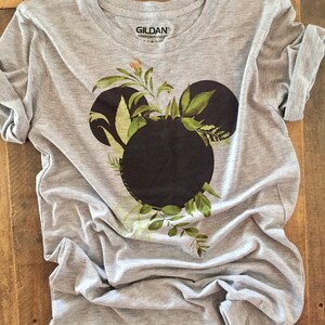 May include: Gray t-shirt with a black Mickey Mouse head silhouette surrounded by green leaves and flowers. The shirt has the Gildan Performance logo on the tag.