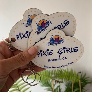 May include: A set of three heart-shaped keychains with a white background and a blue and purple Disneyland castle graphic. The keychains feature the text "Pixie Girls" and "Modesto, CA" with the tagline "Where Dreams Come True".