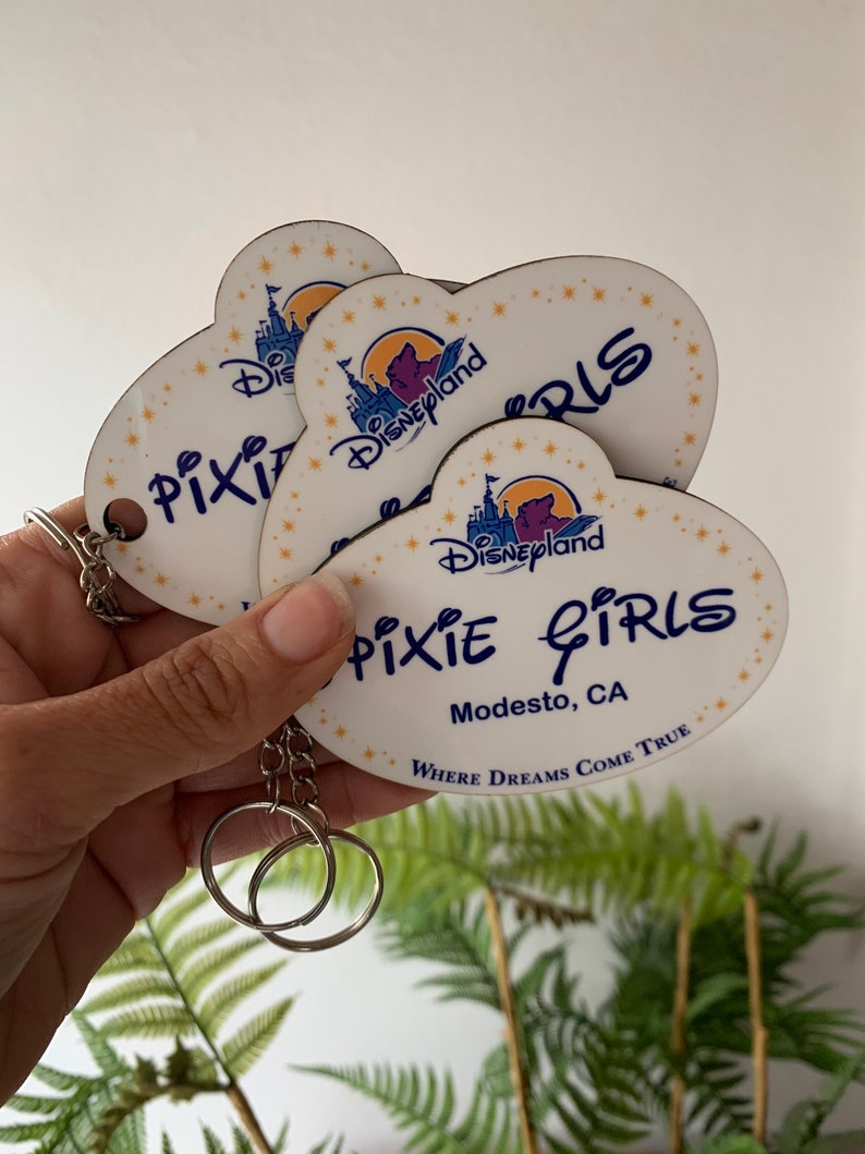 May include: A set of three heart-shaped keychains with a white background and a blue and purple Disneyland castle graphic. The keychains feature the text "Pixie Girls" and "Modesto, CA" with the tagline "Where Dreams Come True".