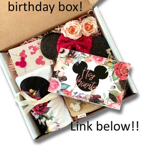 May include: A white gift box filled with a birthday gift set. The box contains a black and white headband with a pink bow, a white t-shirt with a floral print, a card with the text "Hey Beauty" and a floral design, and a small brown pouch with a Mickey Mouse design.