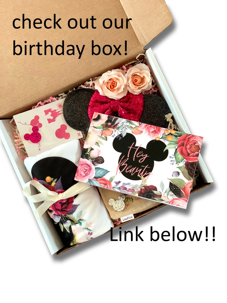 May include: A white gift box filled with a birthday gift set. The box contains a black and white headband with a pink bow, a white t-shirt with a floral print, a card with the text "Hey Beauty" and a floral design, and a small brown pouch with a Mickey Mouse design.