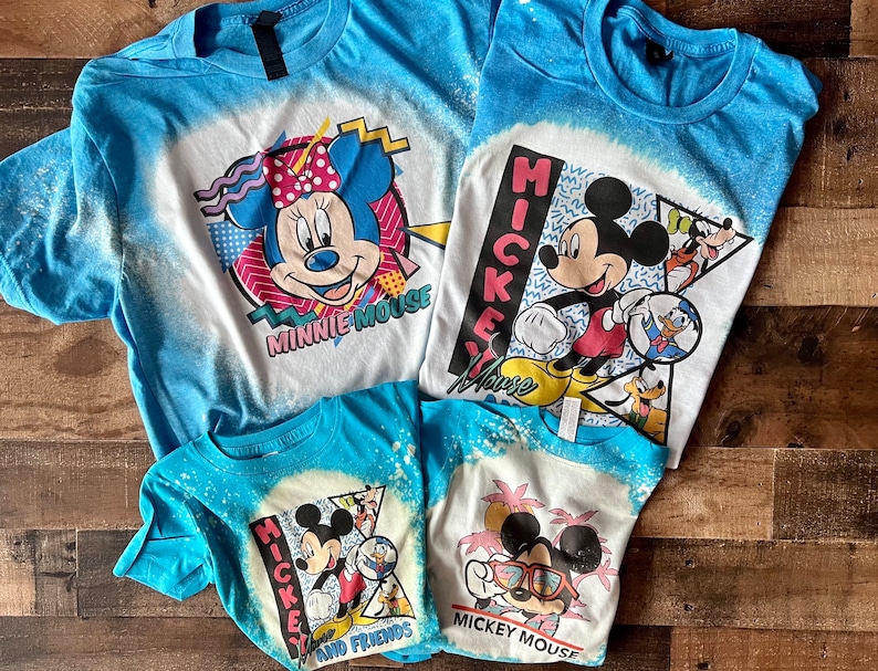 Puede incluir: Cuatro camisetas azules con dise&ntilde;os salpicados de lej&iacute;a con Mickey Mouse y Minnie Mouse. Las camisetas tienen diferentes dise&ntilde;os, incluyendo Mickey Mouse y sus amigos, Mickey Mouse con gafas de sol, Minnie Mouse y Mickey Mouse con Donald Duck y Goofy.