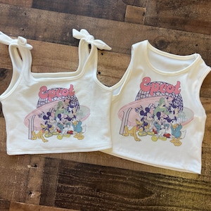 May include: Two white tank tops with the word "Epcot" in pink and a graphic of Disney characters. One top has tie straps, the other has a standard tank top design. Both are on a wooden surface.