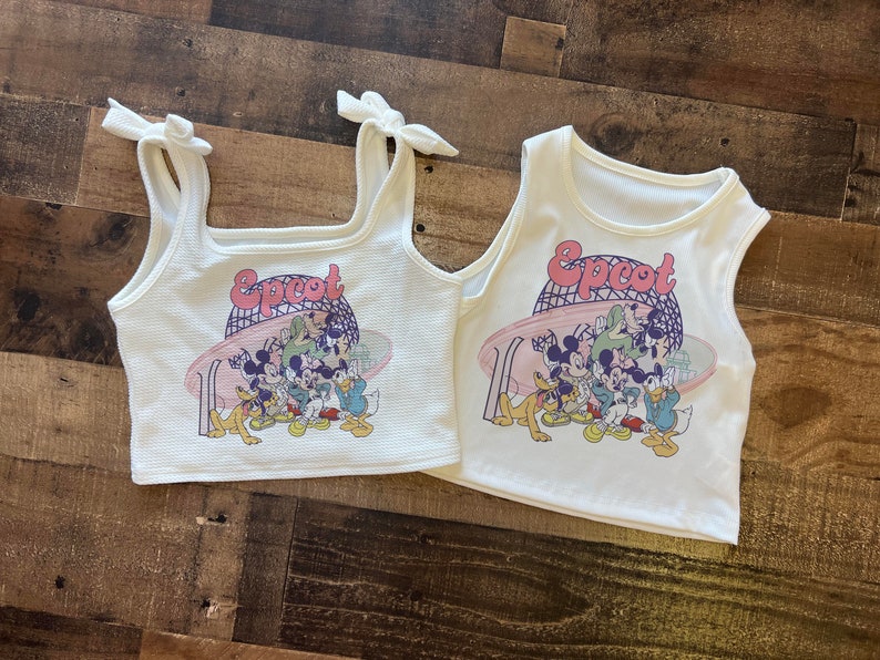 May include: Two white tank tops with the word "Epcot" in pink and a graphic of Disney characters. One top has tie straps, the other has a standard tank top design. Both are on a wooden surface.