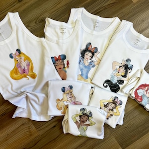 K&ouml;nnte beinhalten: Eine Sammlung wei&szlig;er Tanktops und T-Shirts mit verschiedenen illustrierten weiblichen Charakteren, die Mausohr-Haarreifen tragen. Jedes Kleidungsst&uuml;ck zeigt eine andere Figur mit individuellen Outfits, die mittig auf der Vorderseite aufgedruckt sind.