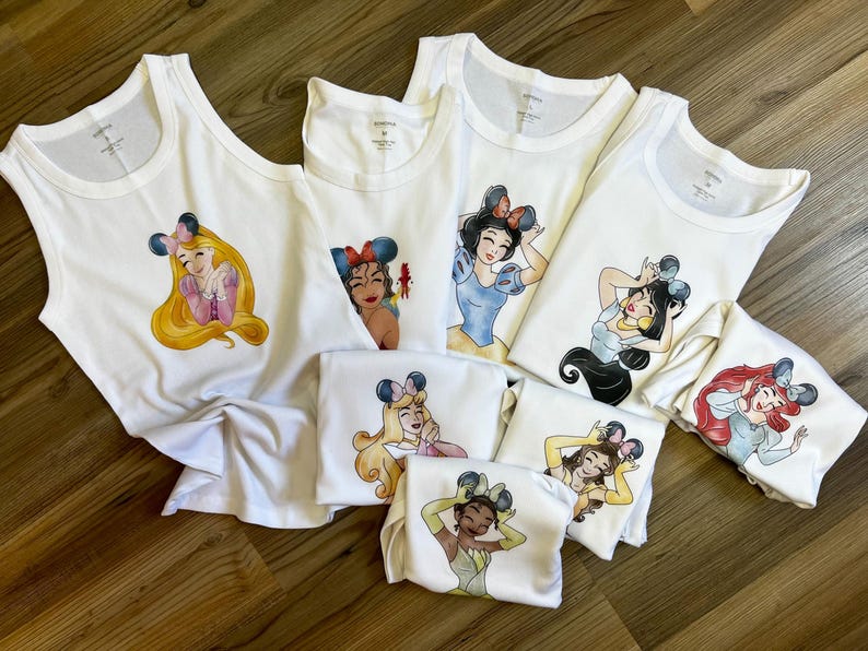 K&ouml;nnte beinhalten: Eine Sammlung wei&szlig;er Tanktops und T-Shirts mit verschiedenen illustrierten weiblichen Charakteren, die Mausohr-Haarreifen tragen. Jedes Kleidungsst&uuml;ck zeigt eine andere Figur mit individuellen Outfits, die mittig auf der Vorderseite aufgedruckt sind.